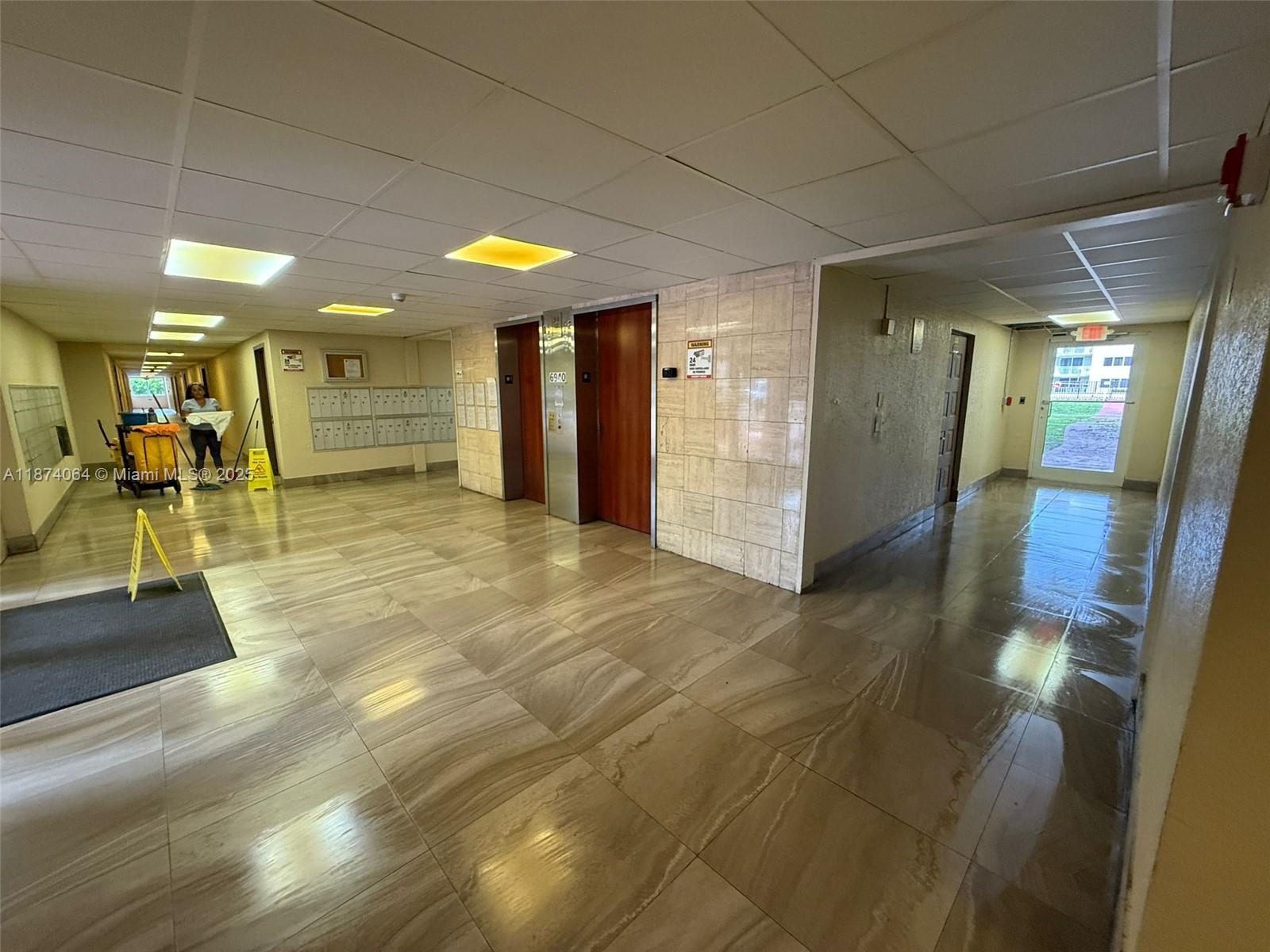 A tiled apartment building lobby features elevators, mailboxes, and a person cleaning the floor — 6940 Miami Gardens Dr # 122, Hialeah FL 33015, HIALEAH, FL