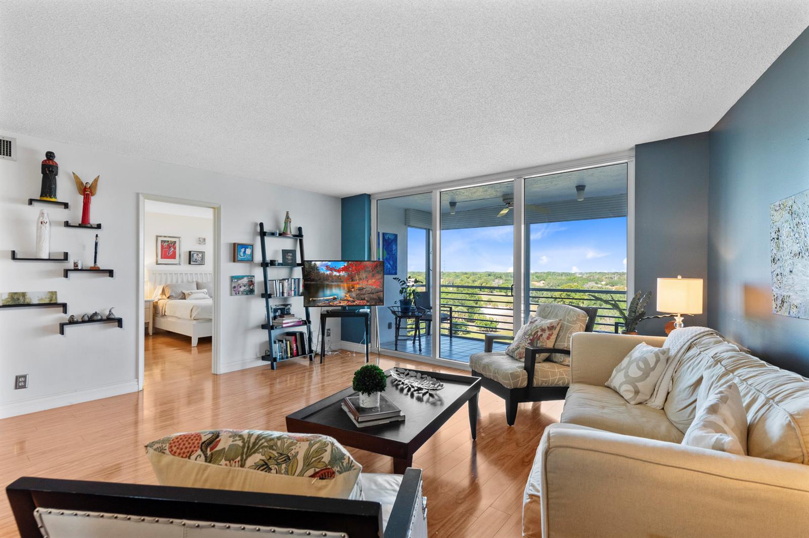 A living room with hardwood floors, a balcony view, and a sofa facing a television — 3499 Oaks Way # 806, Pompano Beach FL 33069, POMPANO BEACH, FL