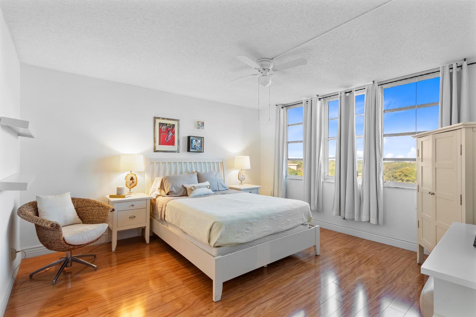 A bedroom with a white bed, two nightstands, a wicker chair, and large windows — 3499 Oaks Way # 806, Pompano Beach FL 33069, POMPANO BEACH, FL