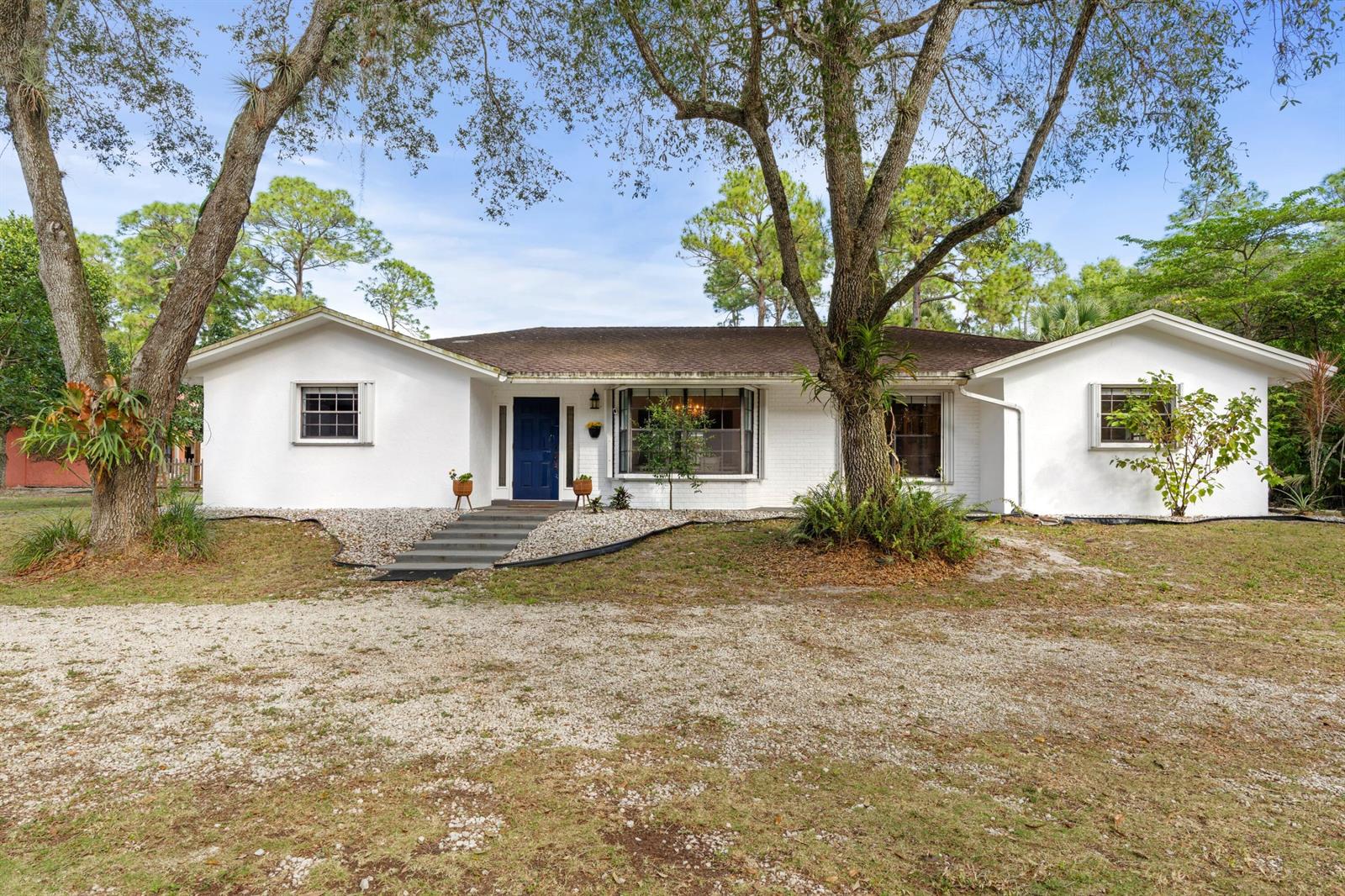 A white, single-story house with a blue front door and a gravel driveway — 14817 Snail Trail, FL 33470, FL