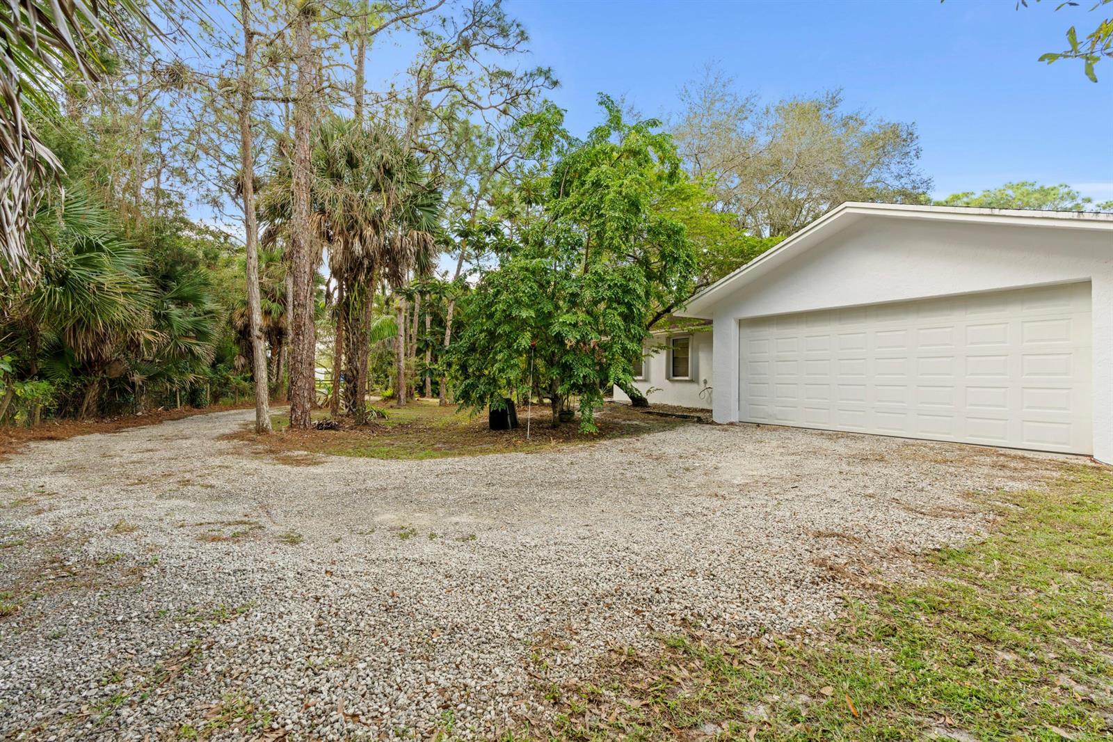 A gravel driveway leads to a white house with a two-car garage and surrounding trees — 14817 Snail Trail, FL 33470, FL