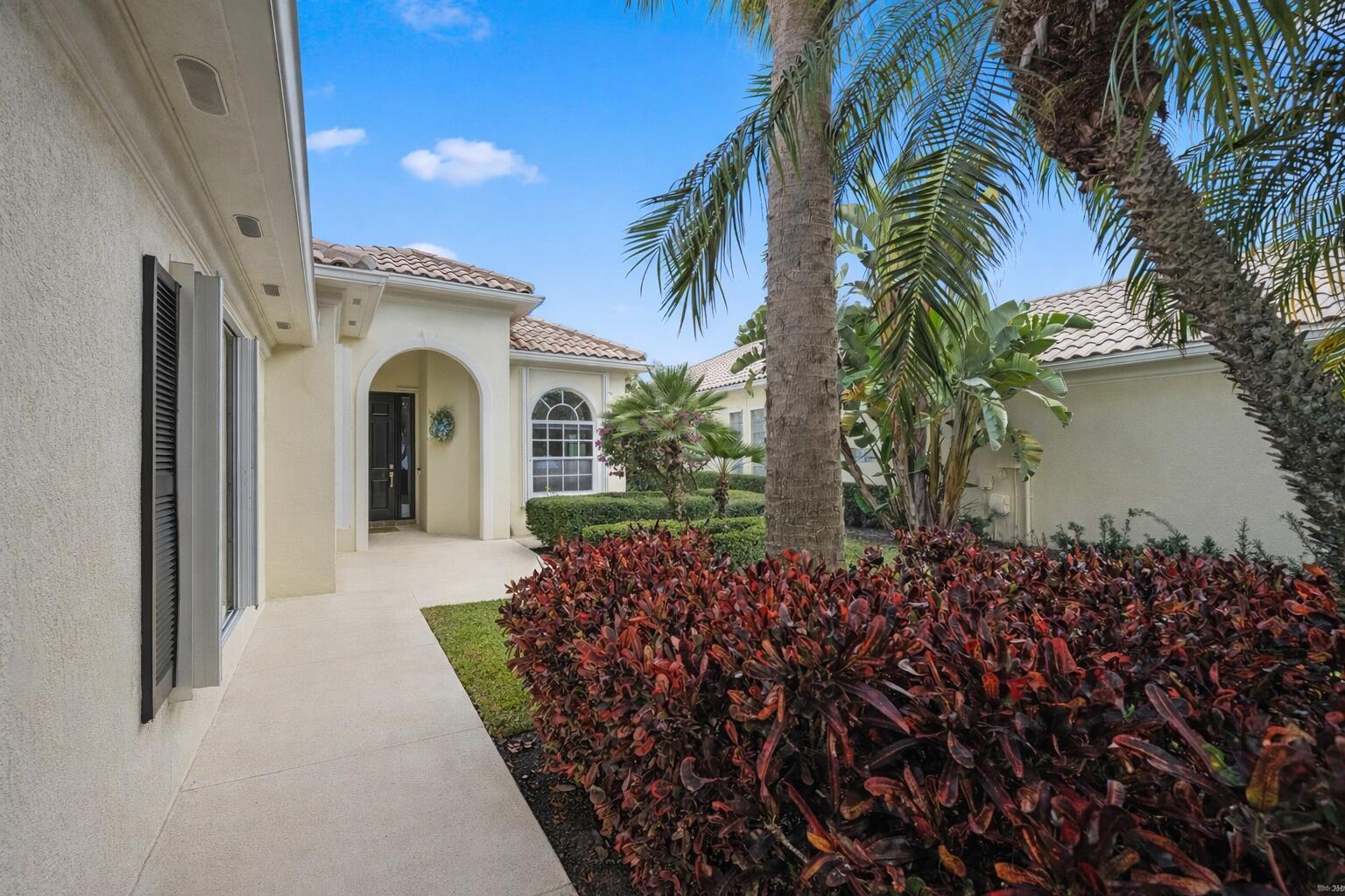 A concrete walkway leads to the arched entryway of a house with tropical landscaping — 8049 Nevis Place, Wellington FL 33414, WELLINGTON, FL