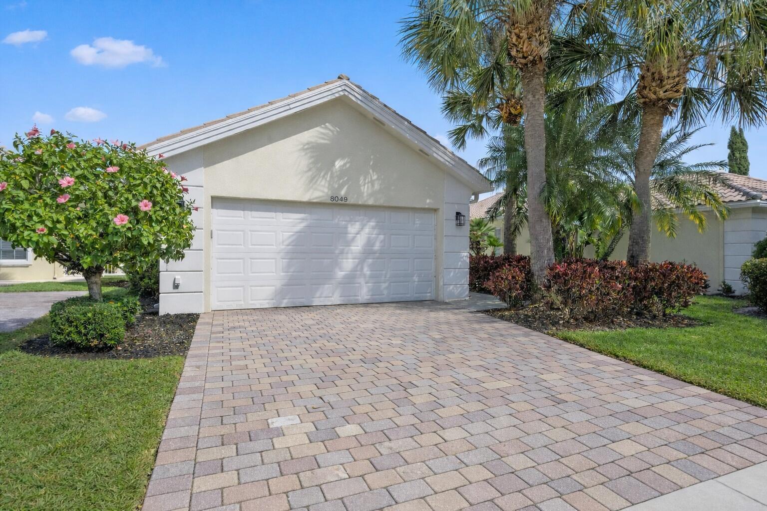 A single-story house exterior featuring a white garage door, paver driveway, and palm trees — 8049 Nevis Place, Wellington FL 33414, WELLINGTON, FL