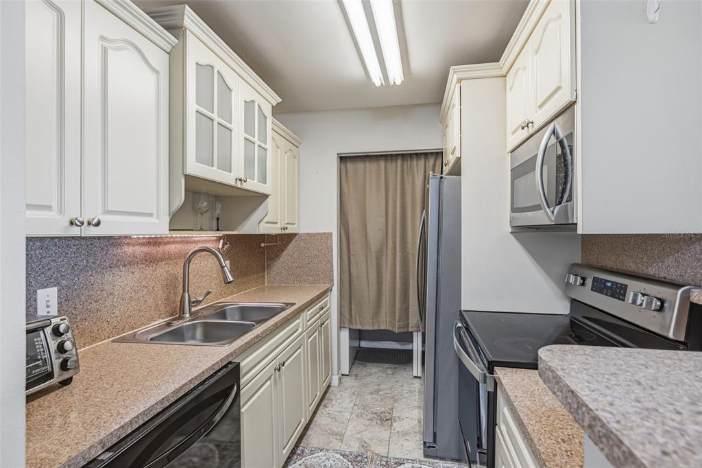 A galley kitchen features off-white cabinets, speckled countertops, stainless steel appliances, and a curtained doorway — 455 ALT 19 S UNIT 163, PALM HARBOR, FL