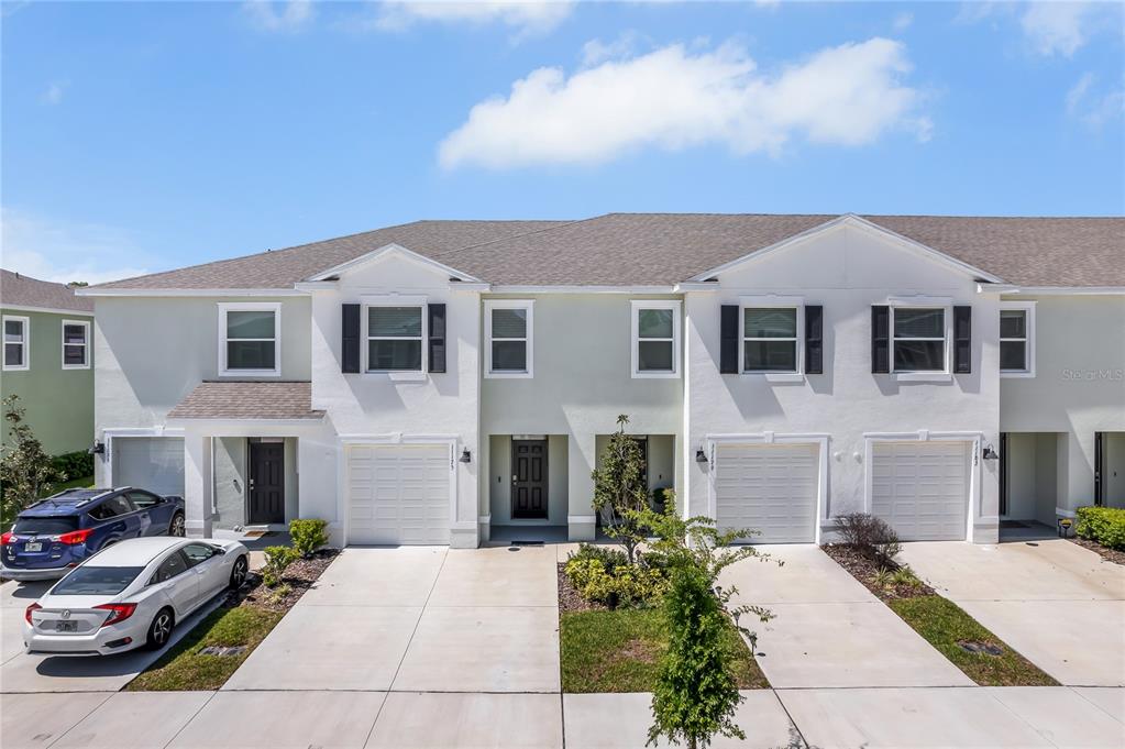 A row of multi-story townhomes features white stucco facades, dark shutters, and attached garages — 11175 CRESCENT DEER DRIVE, LAND O LAKES, FL