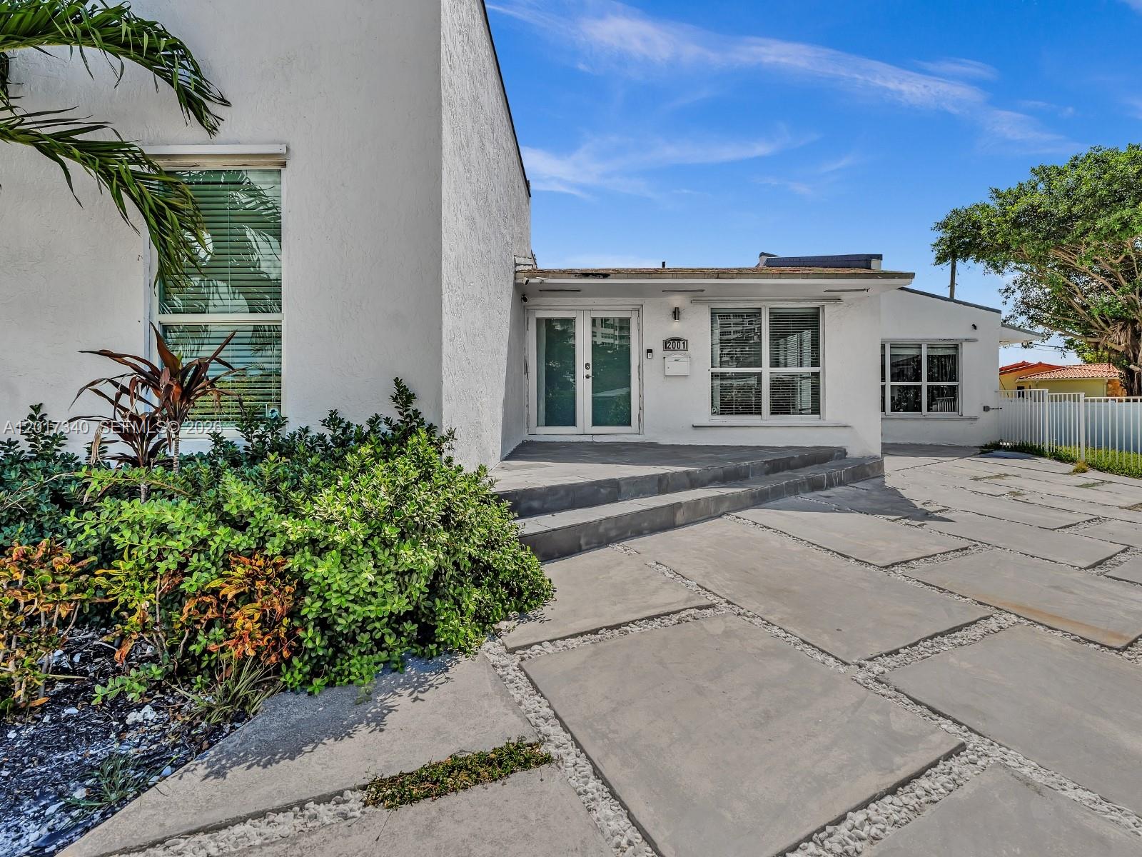 A white house exterior features a paved driveway, a front patio, and lush landscaping — 2001 NE 122nd Rd, North Miami FL 33181, NORTH MIAMI, FL