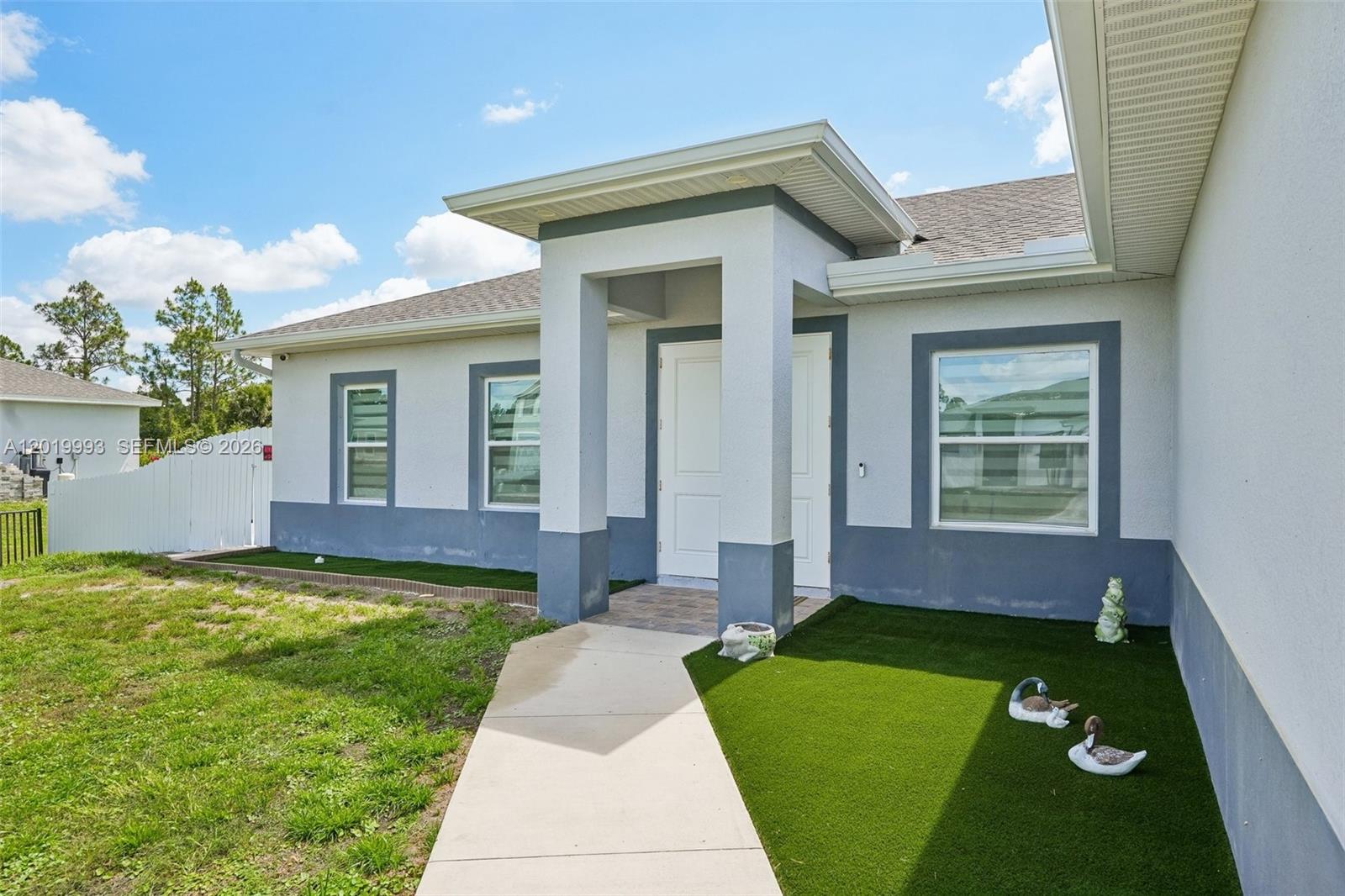 The exterior of a single-story house features a covered entryway, white stucco walls, and artificial turf — 910 Richmond Ave N, Lehigh Acres FL 33972, LEHIGH ACRES, FL