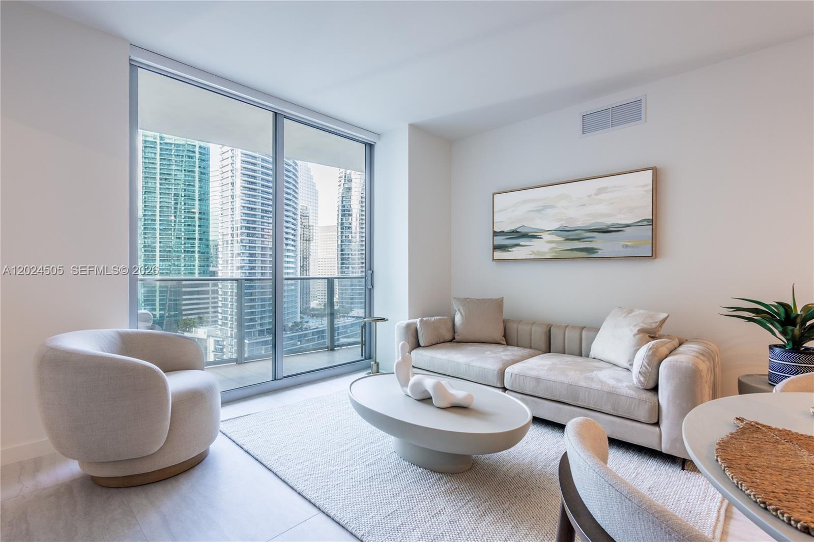 A modern living room features a beige sofa, a round coffee table, and floor-to-ceiling windows — 77 SE 5TH STREET # 2602, Miami FL 33131, MIAMI, FL