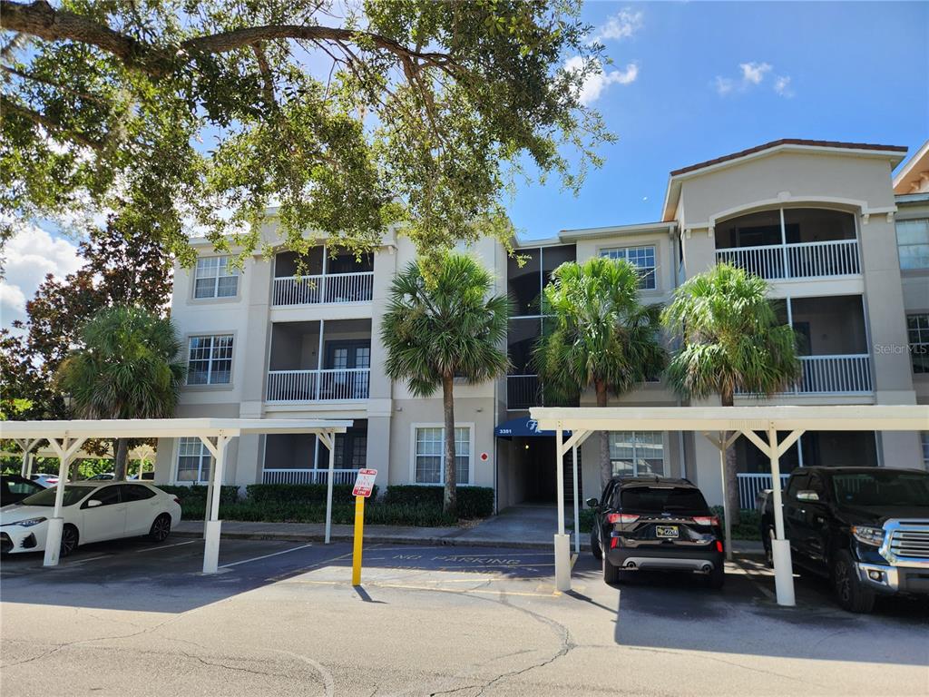 A three-story apartment building features covered parking spaces and palm trees in the foreground — 3351 WHITESTONE CIRCLE UNIT 304, KISSIMMEE, FL