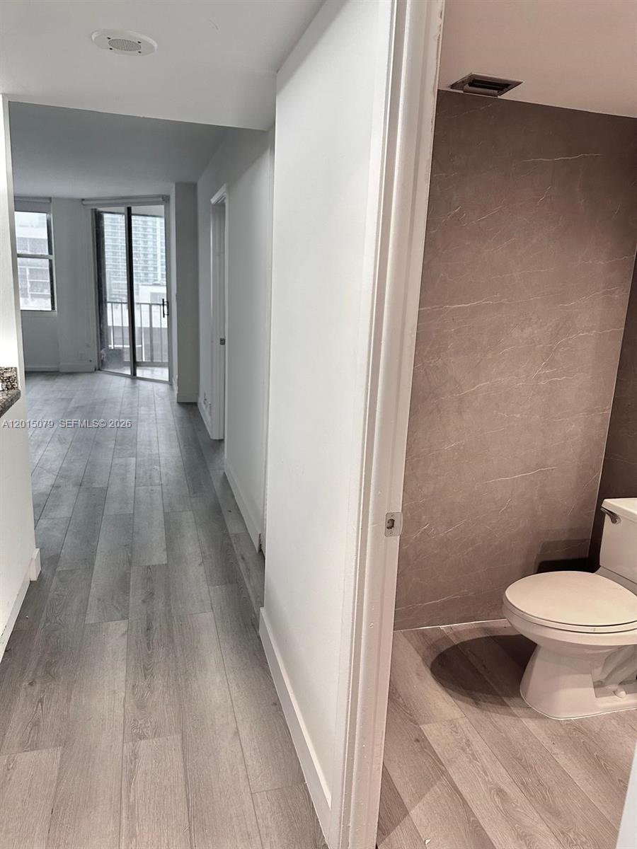 A bathroom with a toilet and tiled wall opens into a hallway with wood-look flooring — 1901 Brickell Ave # B2104, Miami FL 33129, MIAMI, FL