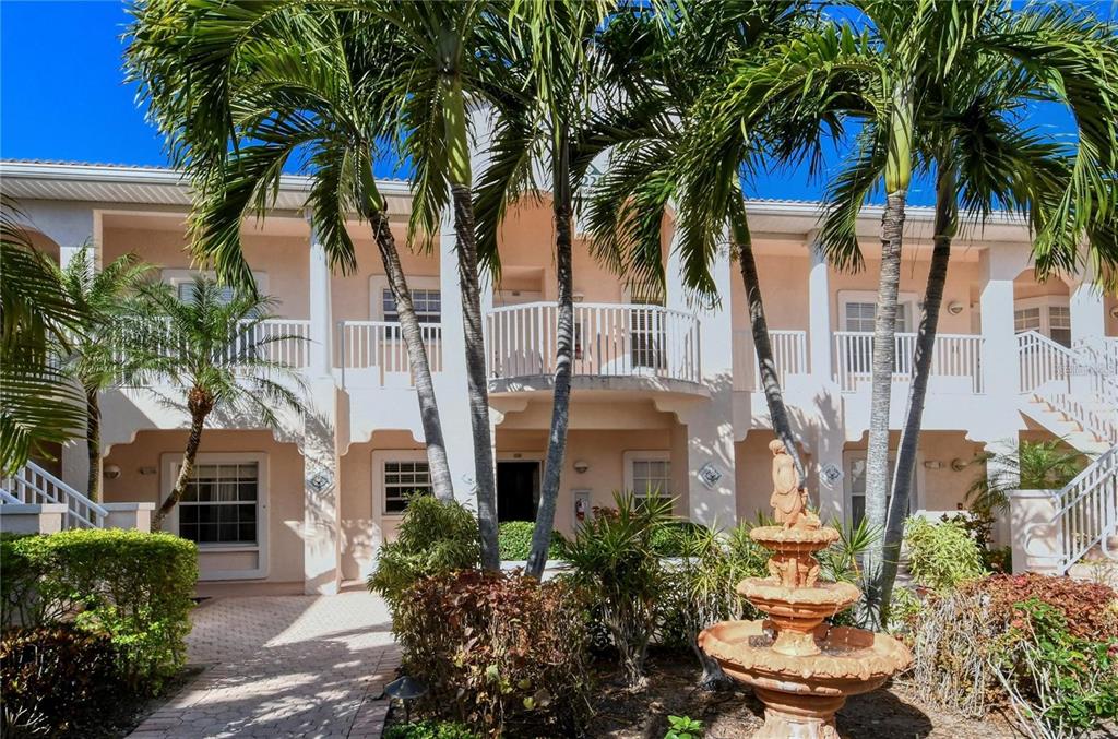 A two-story stucco building with palm trees, a tiered fountain, and balconies under a blue sky — 4224 CENTRAL SARASOTA PARKWAY UNIT 1116, SARASOTA, FL