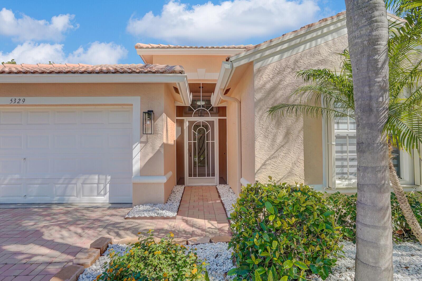 A peach-colored house exterior features a white garage door, a glass storm door, and landscaping — 5329 Toscana Trail, Boynton Beach FL 33437, BOYNTON BEACH, FL