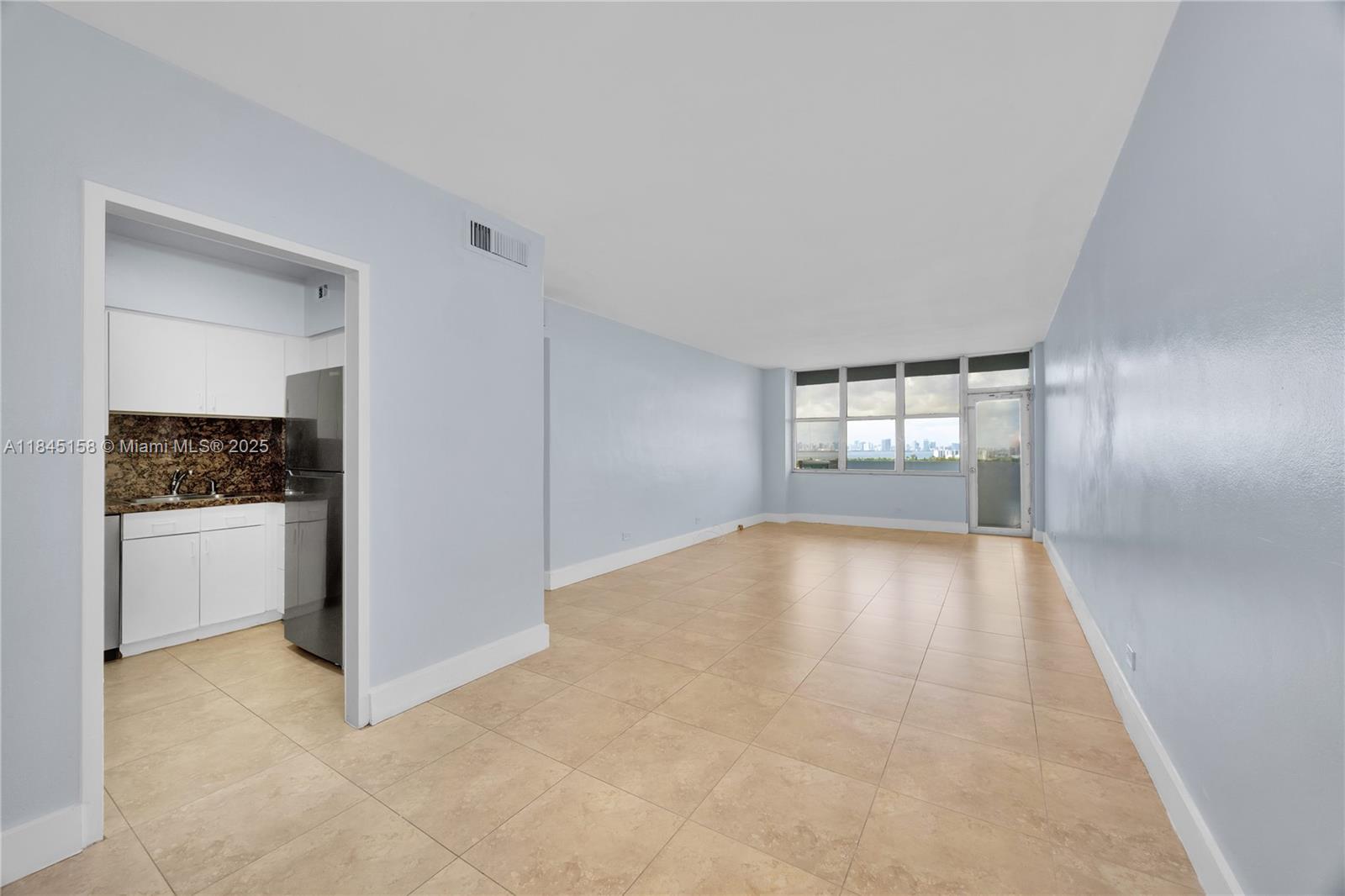 A living area with light blue walls and tile flooring connects to a small kitchen — 7441 Wayne Ave # 15E, Miami Beach FL 33141, MIAMI BEACH, FL