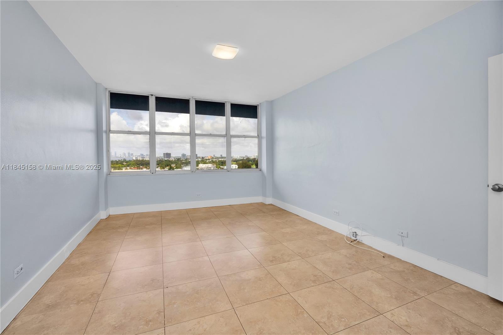 An empty room features light blue walls, tan tile flooring, and large windows with city views — 7441 Wayne Ave # 15E, Miami Beach FL 33141, MIAMI BEACH, FL