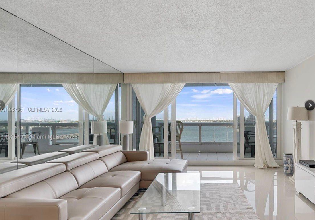 A living room features a beige sectional sofa, glass coffee table, mirrored wall, and balcony view — 1717 N Bayshore Dr # 1132, Miami FL 33132, MIAMI, FL