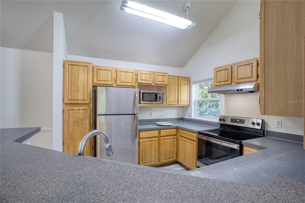A kitchen features light wood cabinets, gray countertops, stainless steel appliances, and a vaulted ceiling — 3433 SOHO STREET UNIT 202, ORLANDO, FL