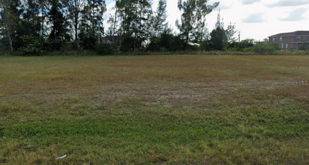 A flat, grassy vacant lot is bordered by trees and a house in the distance — 212 NE 18TH STREET, CAPE CORAL, FL