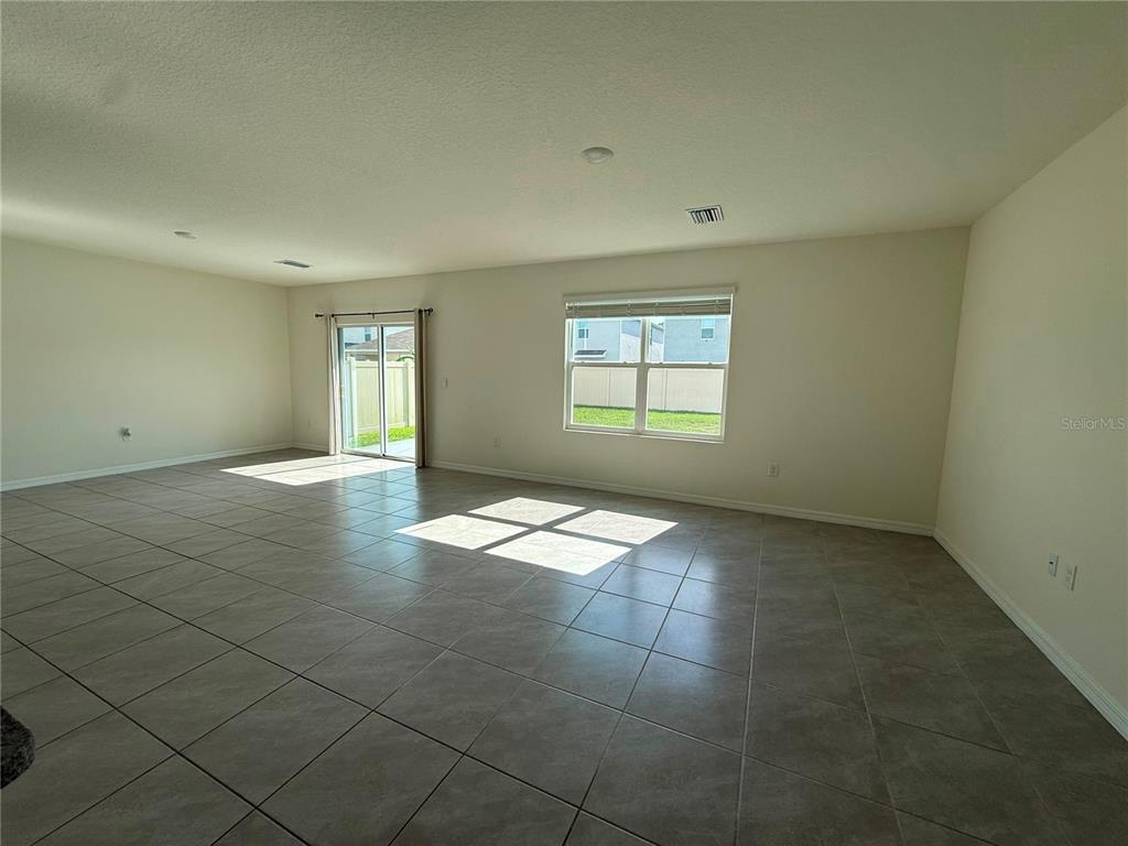A spacious, empty living room features light-colored tile flooring, a sliding glass door, and windows — 2763 COMMON FIG RUN, WESLEY CHAPEL, FL