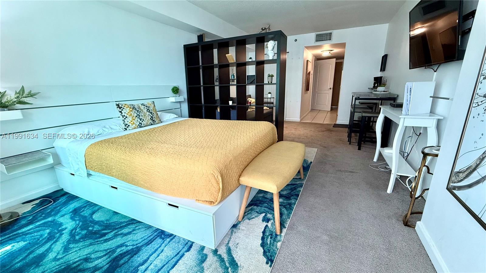 A bedroom features a yellow bed, a dark shelving unit divider, and a blue rug — 1200 Brickell Bay Dr # 2216, Miami FL 33131, MIAMI, FL