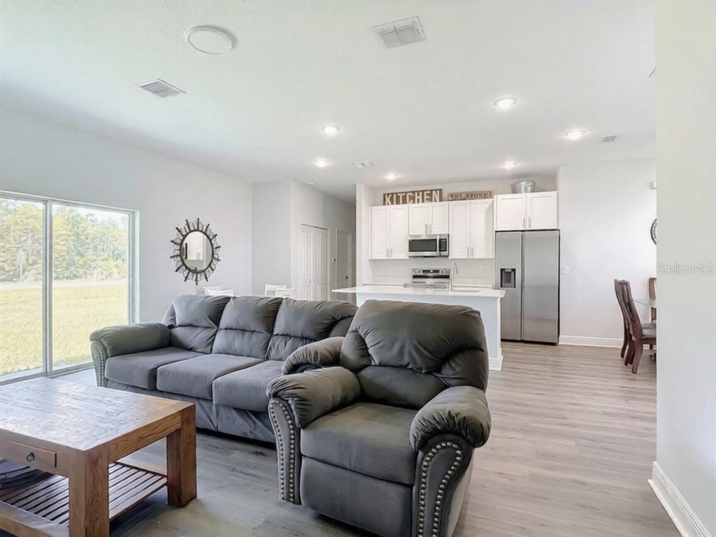 An open-concept living room features gray furniture, wood-look flooring, and a white kitchen in back — 1101 SAINT TROPEZ COURT, KISSIMMEE, FL