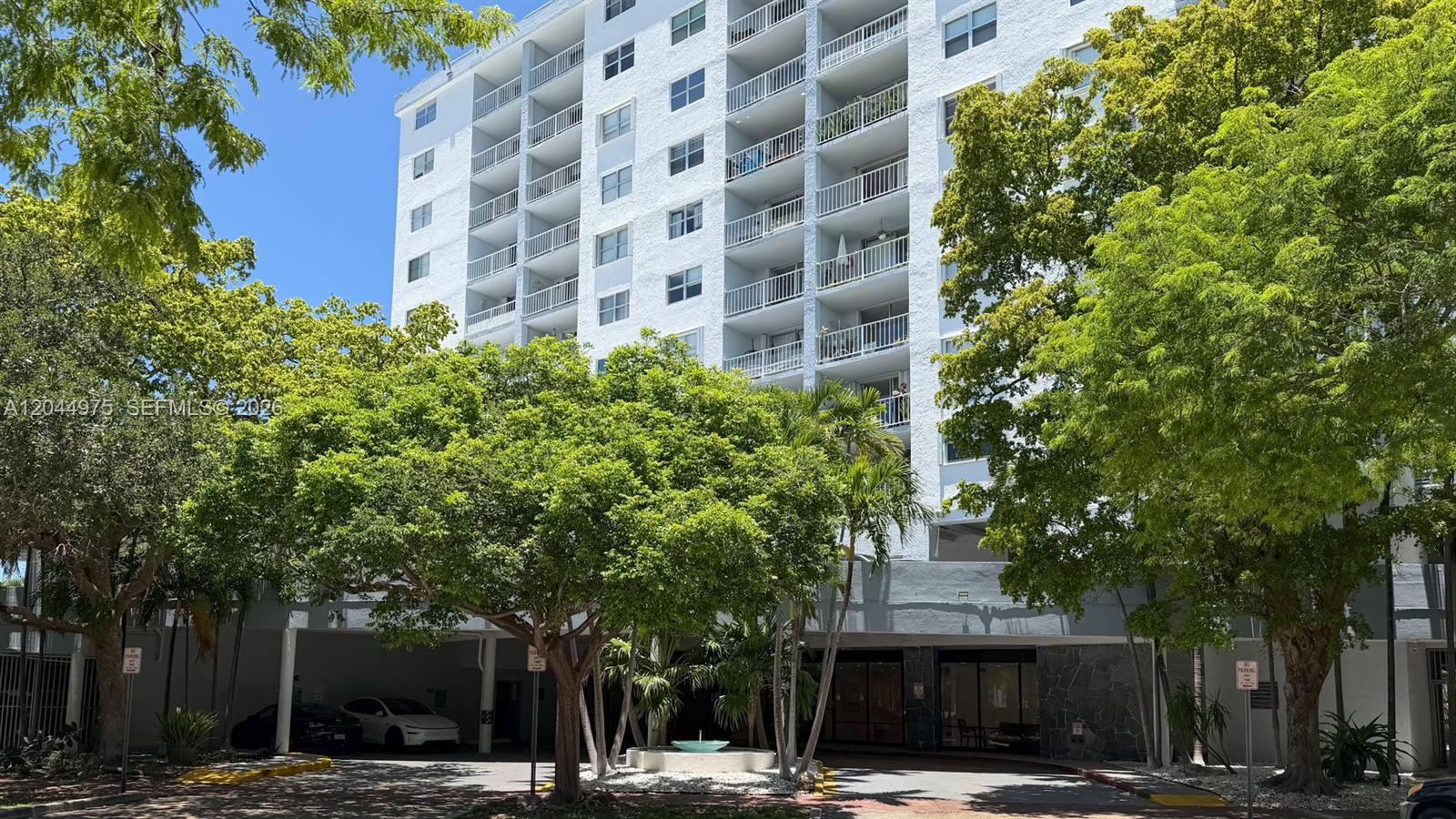 A multi-story white residential building features balconies and a ground-level parking area with trees — 6900 Bay Dr # 6H, Miami Beach FL 33141, MIAMI BEACH, FL