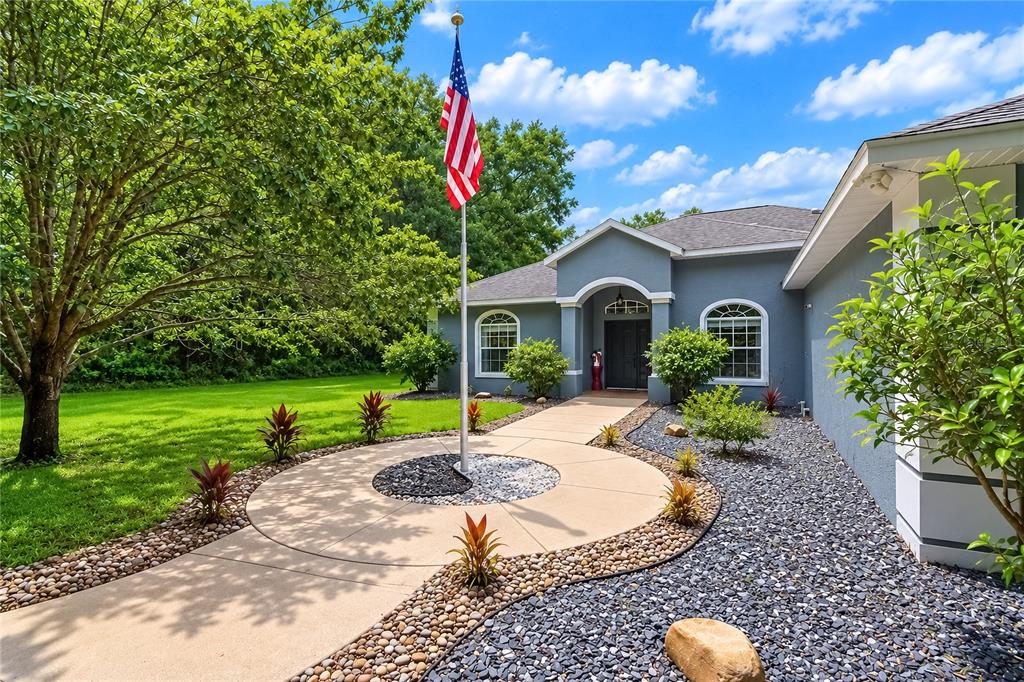 A gray house exterior features a concrete walkway, a flagpole, and decorative rock landscaping — 20130 SW 95TH STREET, DUNNELLON, FL