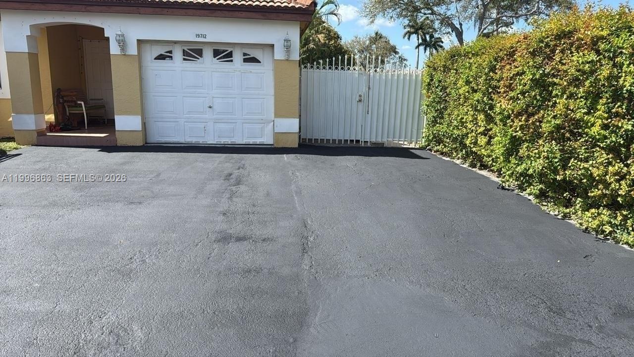 A paved driveway leads to a garage and a white gate beside a tall hedge — 19712 NW 82nd Pl, Hialeah FL 33015, HIALEAH, FL