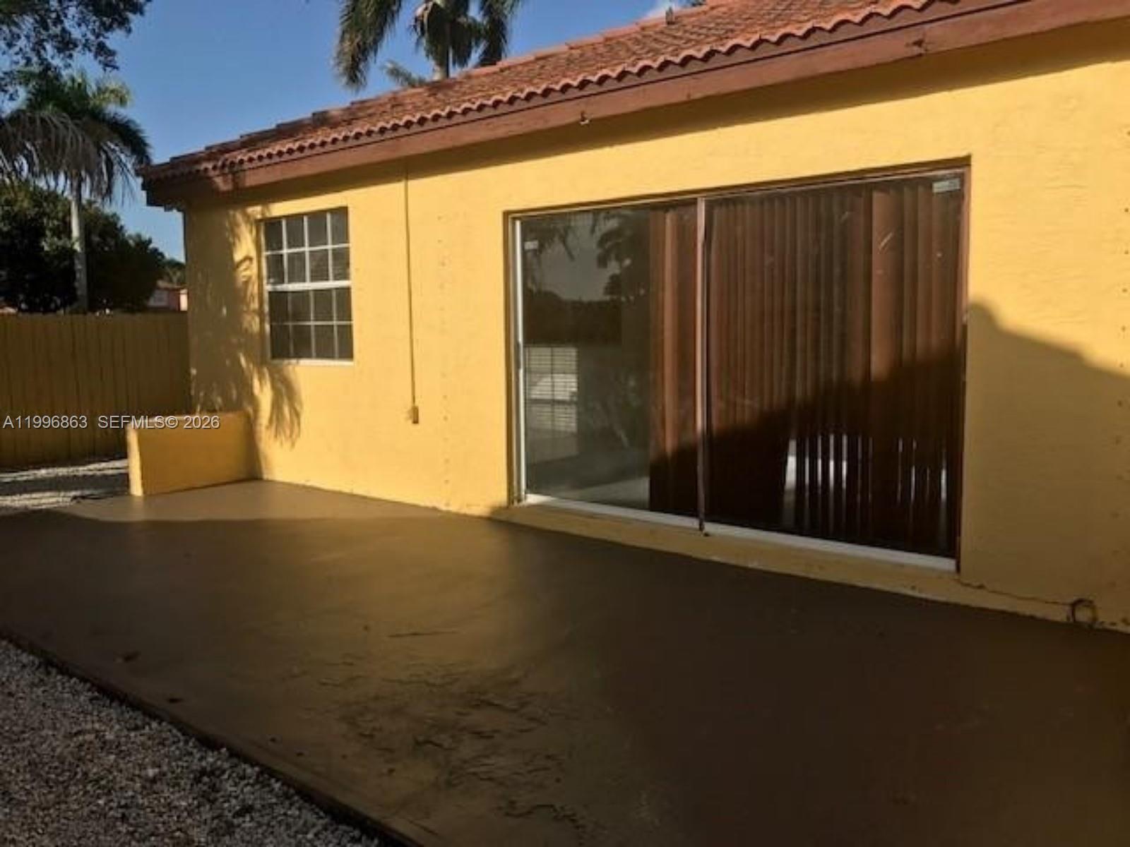 A yellow house exterior features a concrete patio, a sliding glass door, and a window — 19712 NW 82nd Pl, Hialeah FL 33015, HIALEAH, FL
