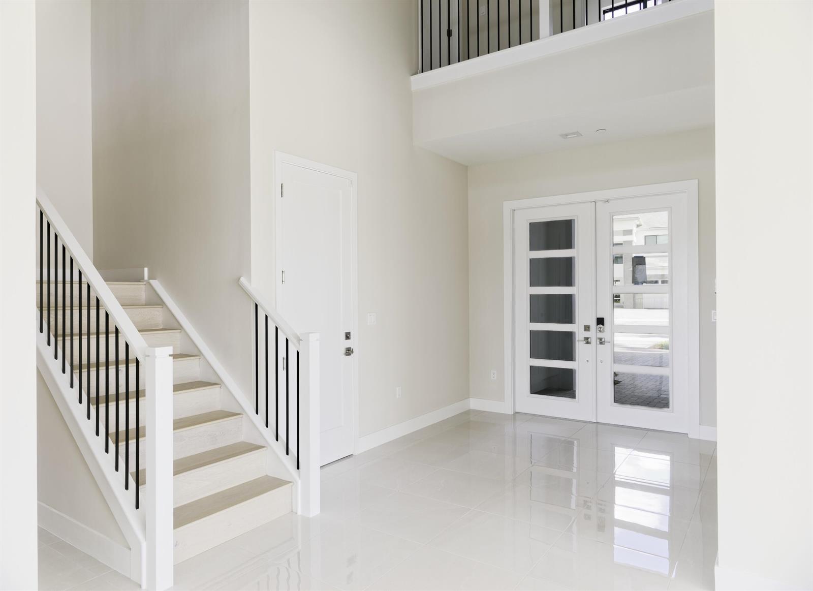 A bright foyer features white tile floors, a staircase with black railings, and double glass doors — 9895 Migration Point, Palm Beach Gardens FL 33412, PALM BEACH GARDENS, FL