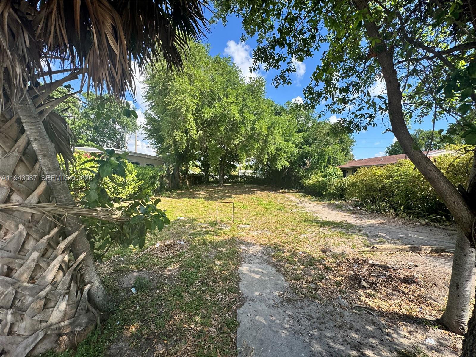 A grassy, partially shaded vacant lot is framed by trees and neighboring residential buildings — 629 NW 65th St, Miami FL 33150, MIAMI, FL