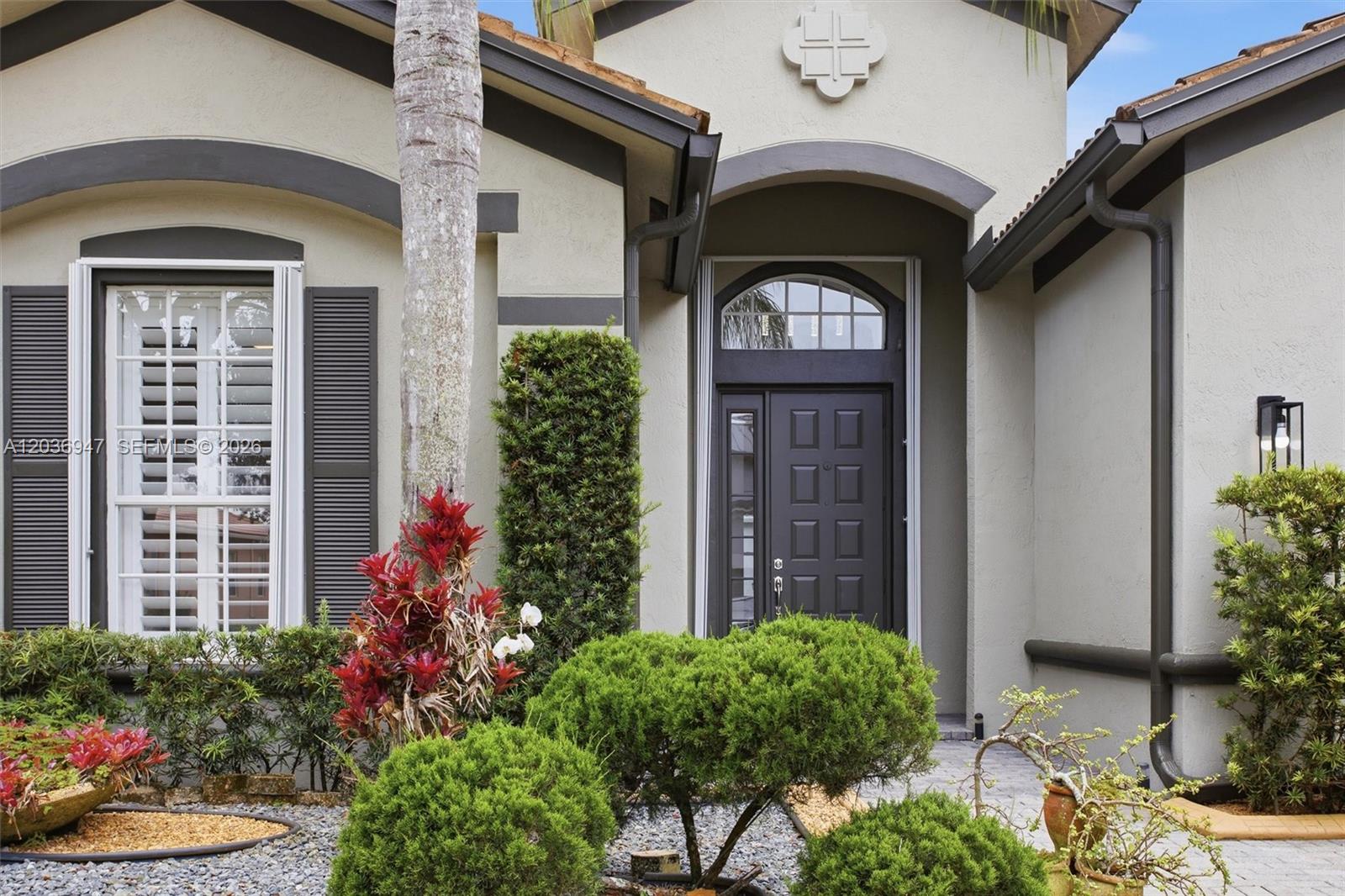 The exterior of a house features a dark front door, arched transom window, and landscaping — 19308 Stonebrook St, Weston FL 33332, WESTON, FL