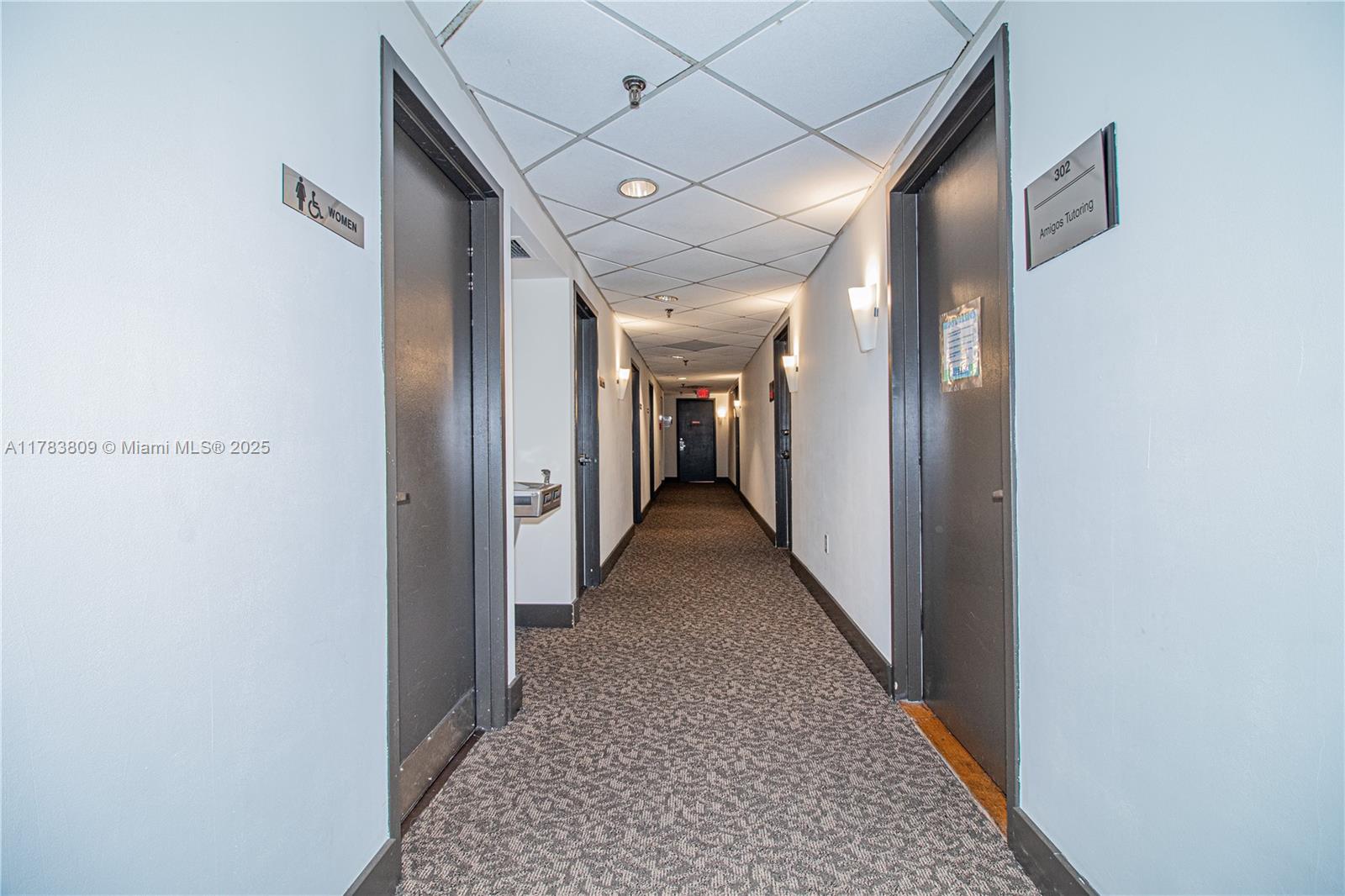 A long, carpeted hallway features several doors, a water fountain, and a drop ceiling — 2100 SW 22nd St # 302, Miami FL 33145, MIAMI, FL