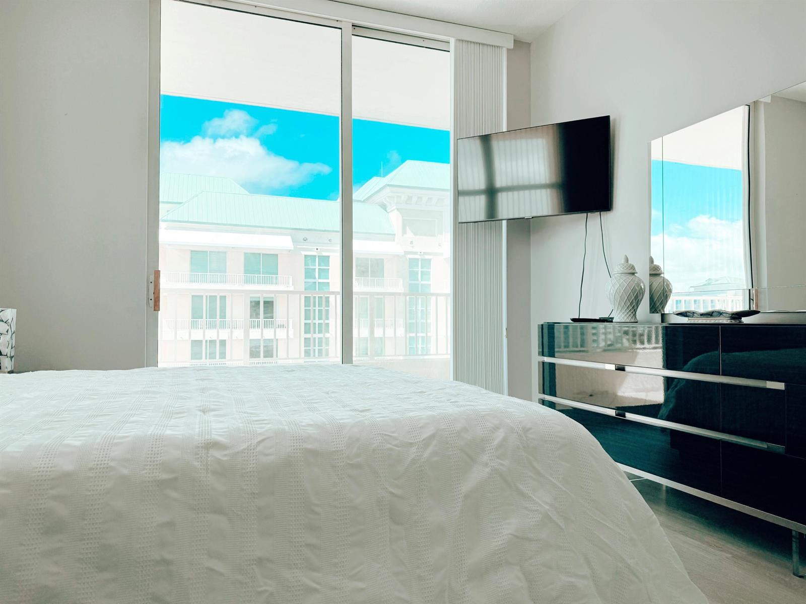 A bedroom features a white bed, a mirrored dresser, a wall-mounted television, and a balcony view — 625 Casa Loma Boulevard Unit 1, Boynton Beach FL 33435, BOYNTON BEACH, FL