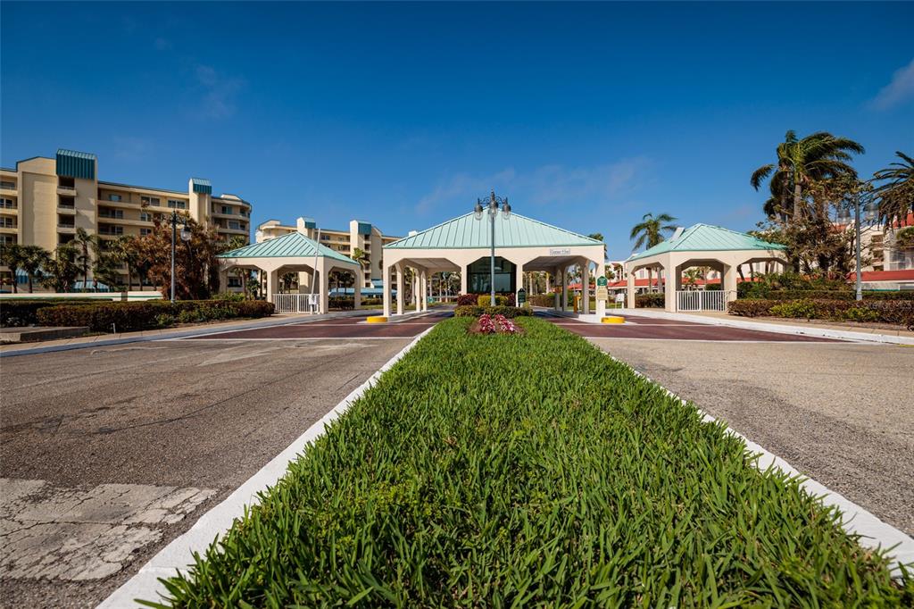 A gated entrance with three covered structures and a central grass median under blue skies — 7897 SAILBOAT KEY BOULEVARD UNIT 107, SOUTH PASADENA, FL