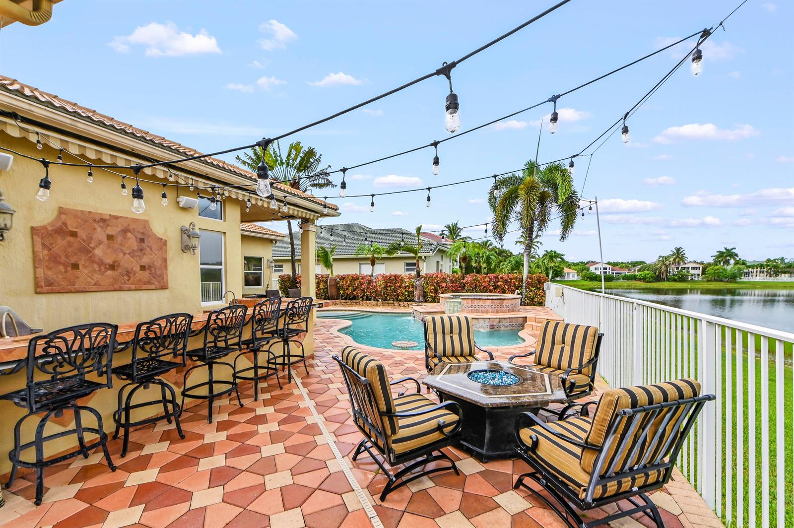 A patio features a bar, fire pit, pool, and string lights overlooking a lake — 11852 Osprey Point Circle, Wellington FL 33449, WELLINGTON, FL