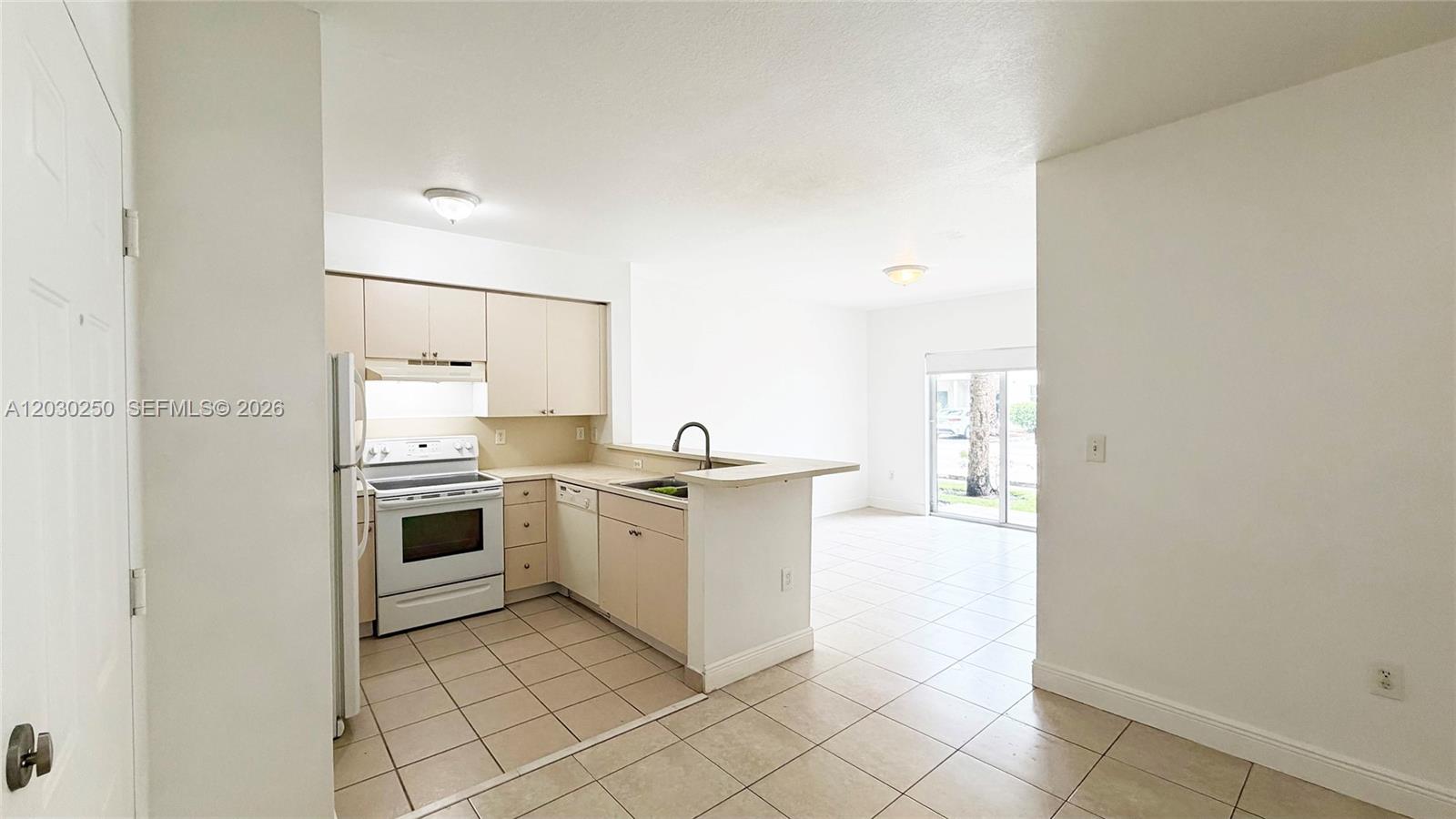 A kitchen with light-colored cabinets and tile flooring opens into a bright living area — CUTLER BAY, FL