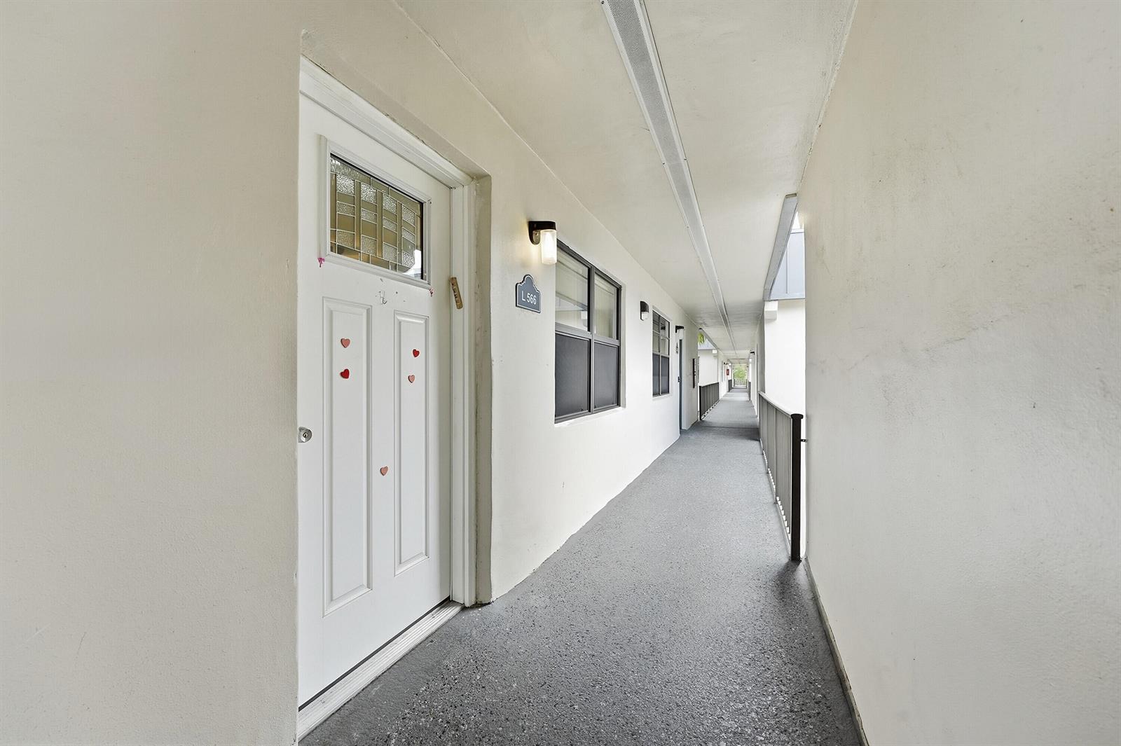 An outdoor apartment hallway features a white door with heart decals and gray flooring — 566 Piedmont L, Delray Beach FL 33484, DELRAY BEACH, FL