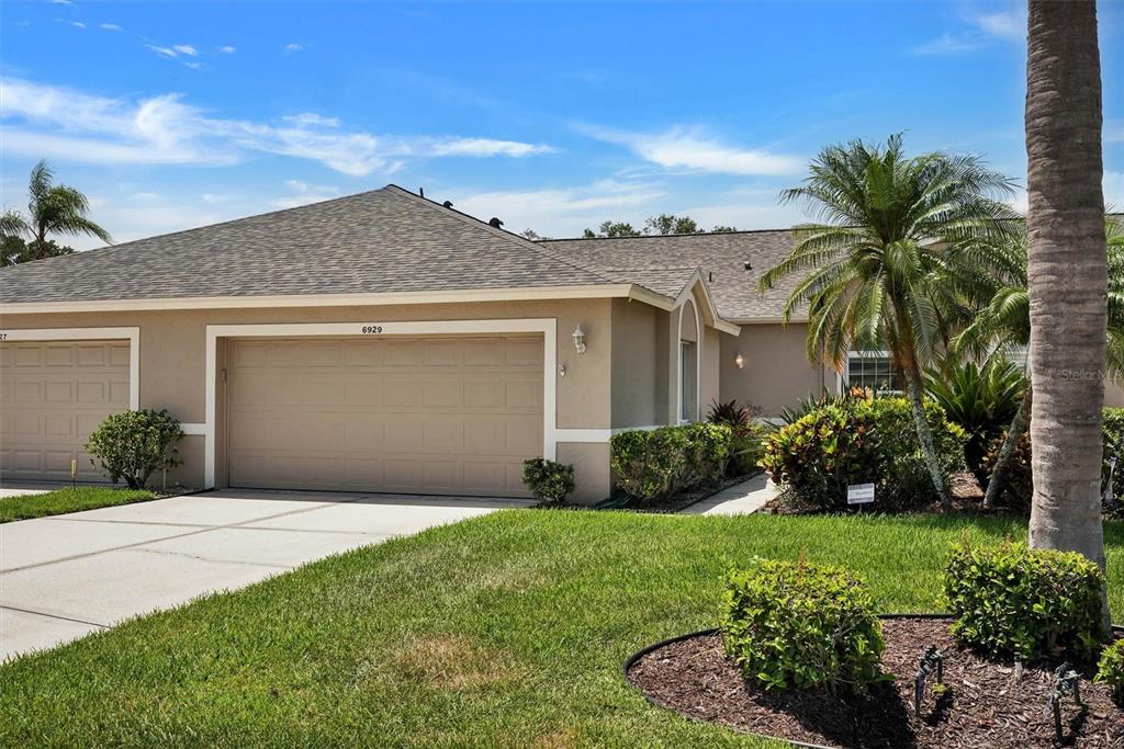 A tan single-story house with a two-car garage, a palm tree, and landscaped front yard — 6929 STONEYWALK COURT, BRADENTON, FL