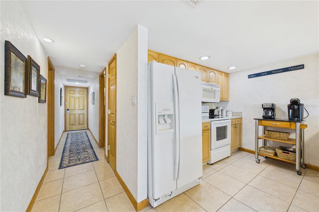 A kitchen with light wood cabinets, white appliances, and tile flooring leads to a hallway — 19610 GULF BOULEVARD UNIT 206, INDIAN ROCKS BEACH, FL