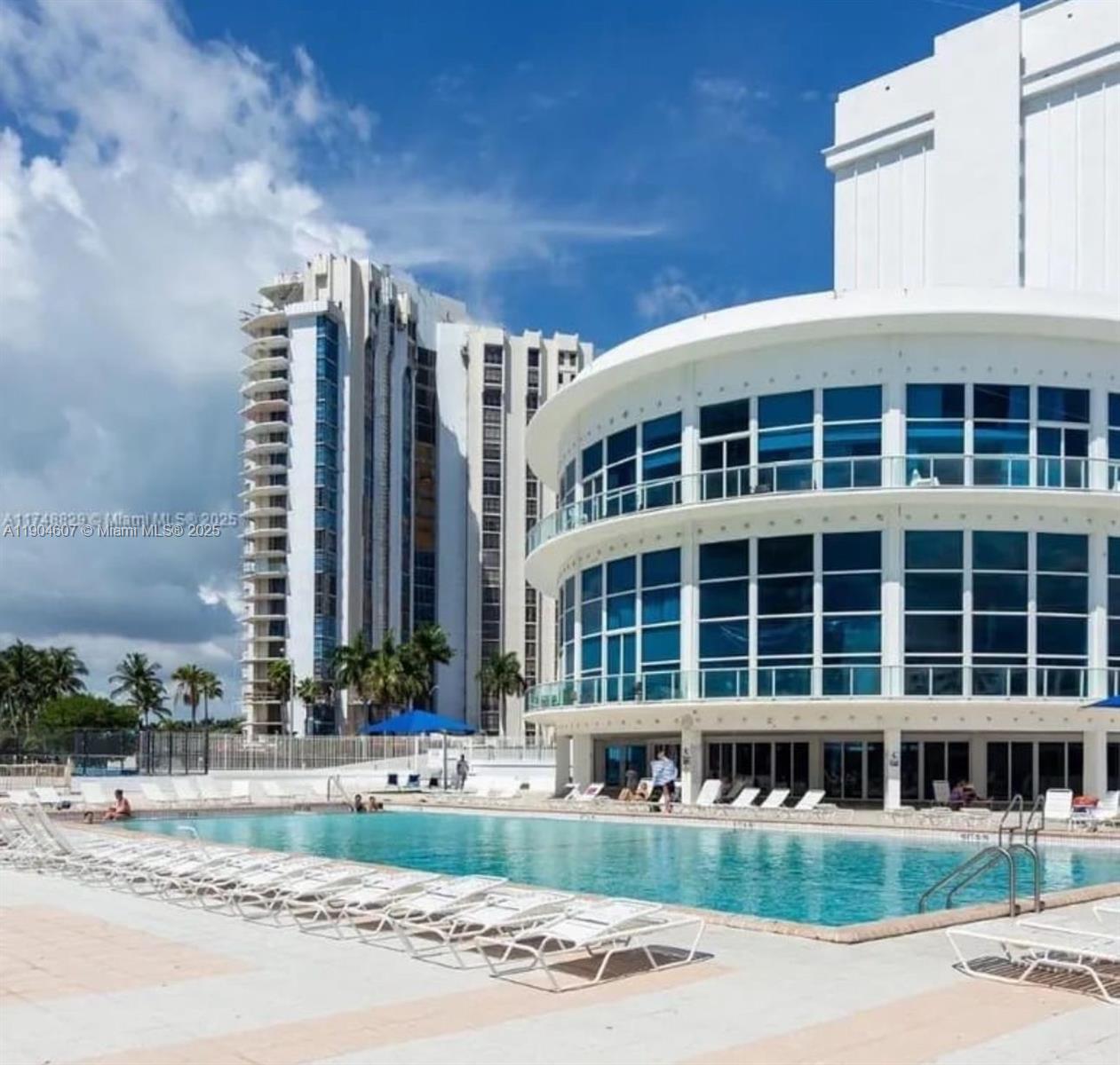 A swimming pool sits in front of a modern white building with large glass windows — 5445 Collins Ave # 617, Miami Beach FL 33140, MIAMI BEACH, FL