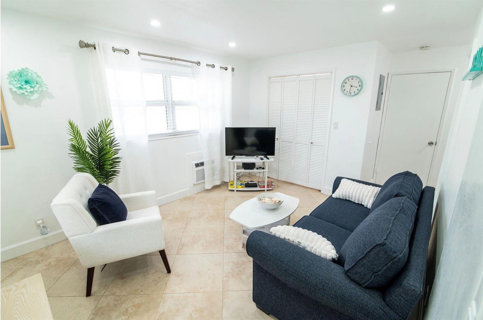 A living room with tile floors, a dark blue sofa, white armchair, and television stand — 3230 NE 13th Street # 203, Pompano Beach FL 33062, POMPANO BEACH, FL