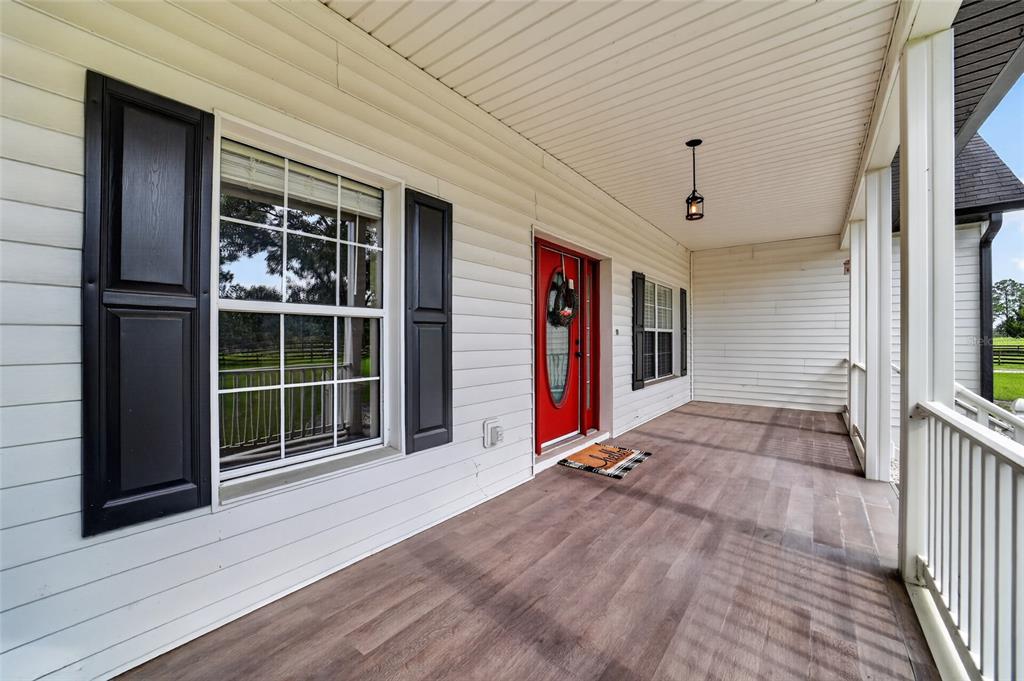 A covered front porch with white siding, black shutters, a red door, and wood-look flooring — 6351 SE 140 AVENUE, MORRISTON, FL