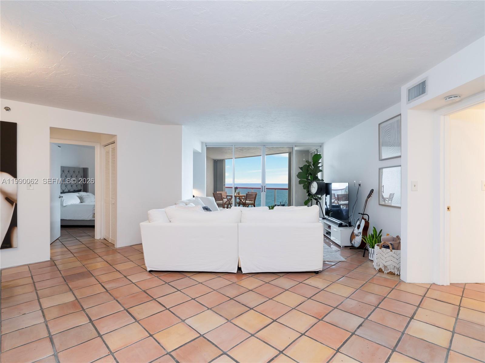 A living room with white furniture and tile floors opens to a balcony overlooking the ocean — 6767 Collins Ave # 1704, Miami Beach FL 33141, MIAMI BEACH, FL