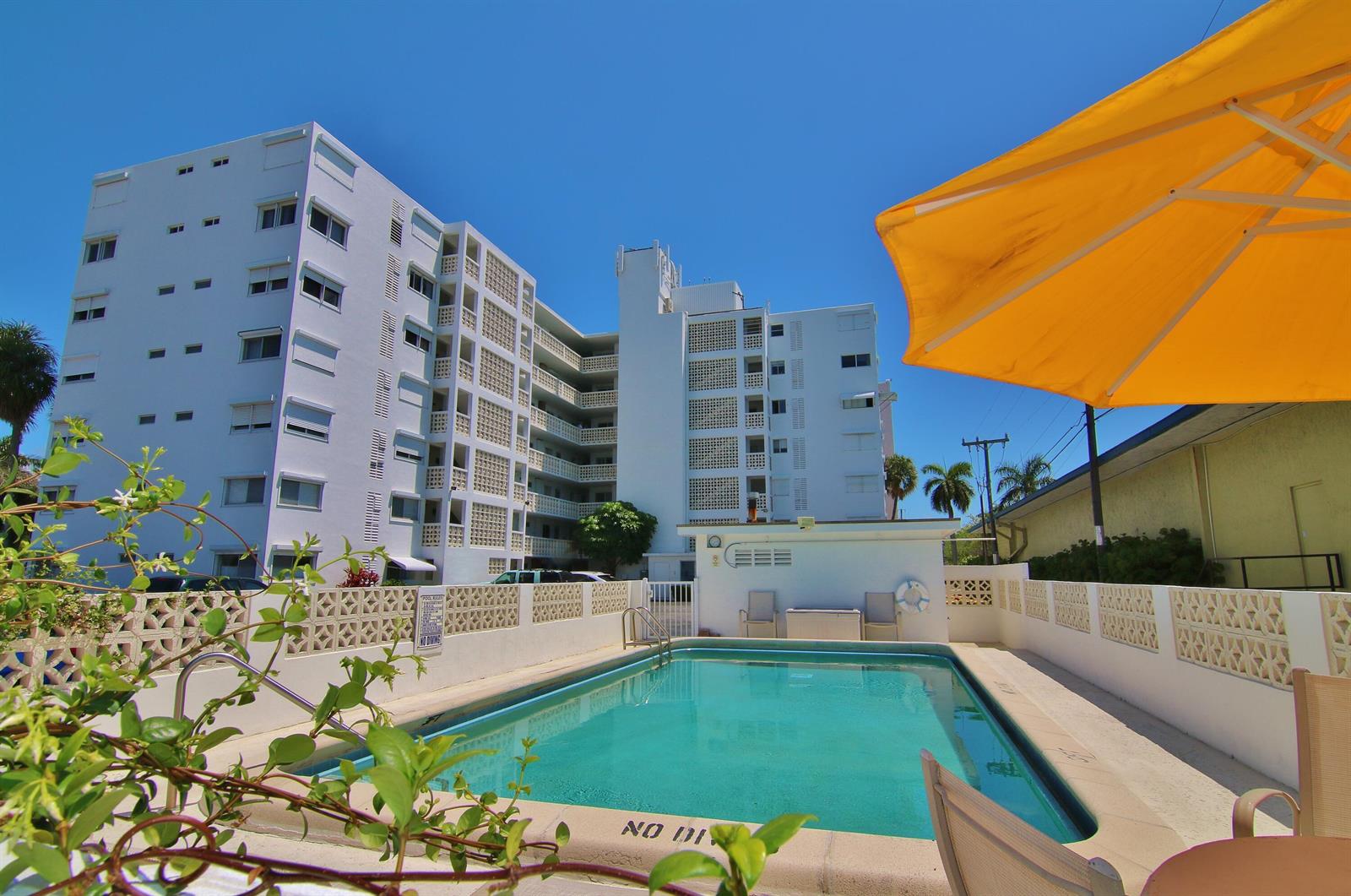 A swimming pool sits in front of a multi-story white apartment building under a blue sky — 1 N Golfview Road # 205, FL 33460, FL
