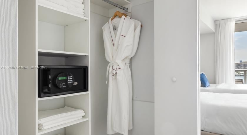 A closet with white shelving, a black safe, and hanging bathrobes next to a bedroom — 2602 E Hallandale Beach Blvd # R310, Hallandale Beach FL 33009, HALLANDALE BEACH, FL