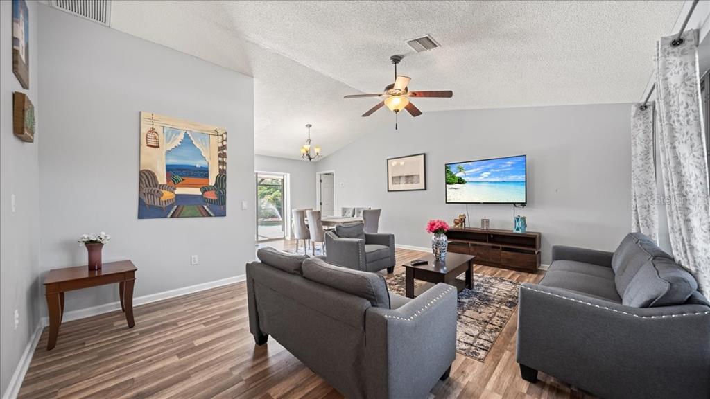 A living room features gray furniture, wood-look flooring, a ceiling fan, and a wall-mounted television — 11195 SEABREEZE AVENUE, PORT CHARLOTTE, FL