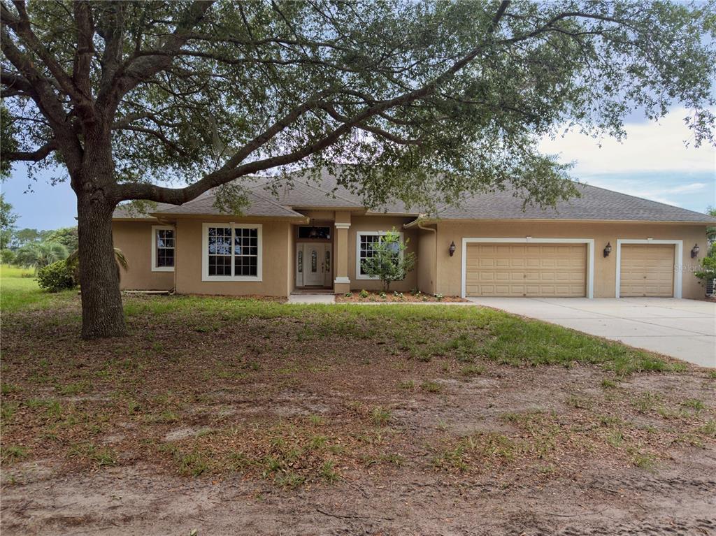 A single-story tan stucco house with a three-car garage and a large oak tree — 24087 TWISTER LANE, BROOKSVILLE, FL