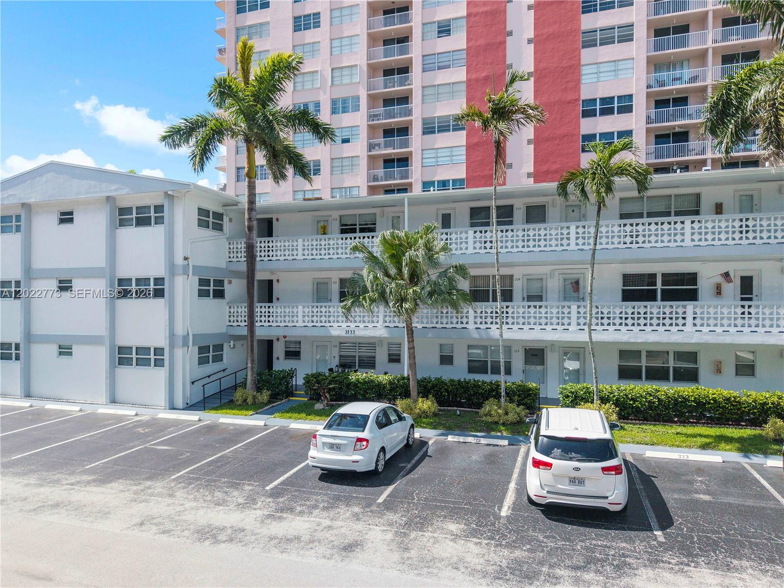 An exterior view of a multi-story apartment building with a parking lot and palm trees — 3133 S Ocean Dr # 318, Hallandale Beach FL 33009, HALLANDALE BEACH, FL