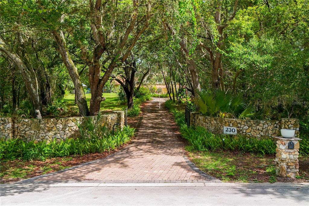 A brick driveway leads through a stone-walled entrance toward a house shaded by large trees — 230 E PANAMA ROAD, WINTER SPRINGS, FL