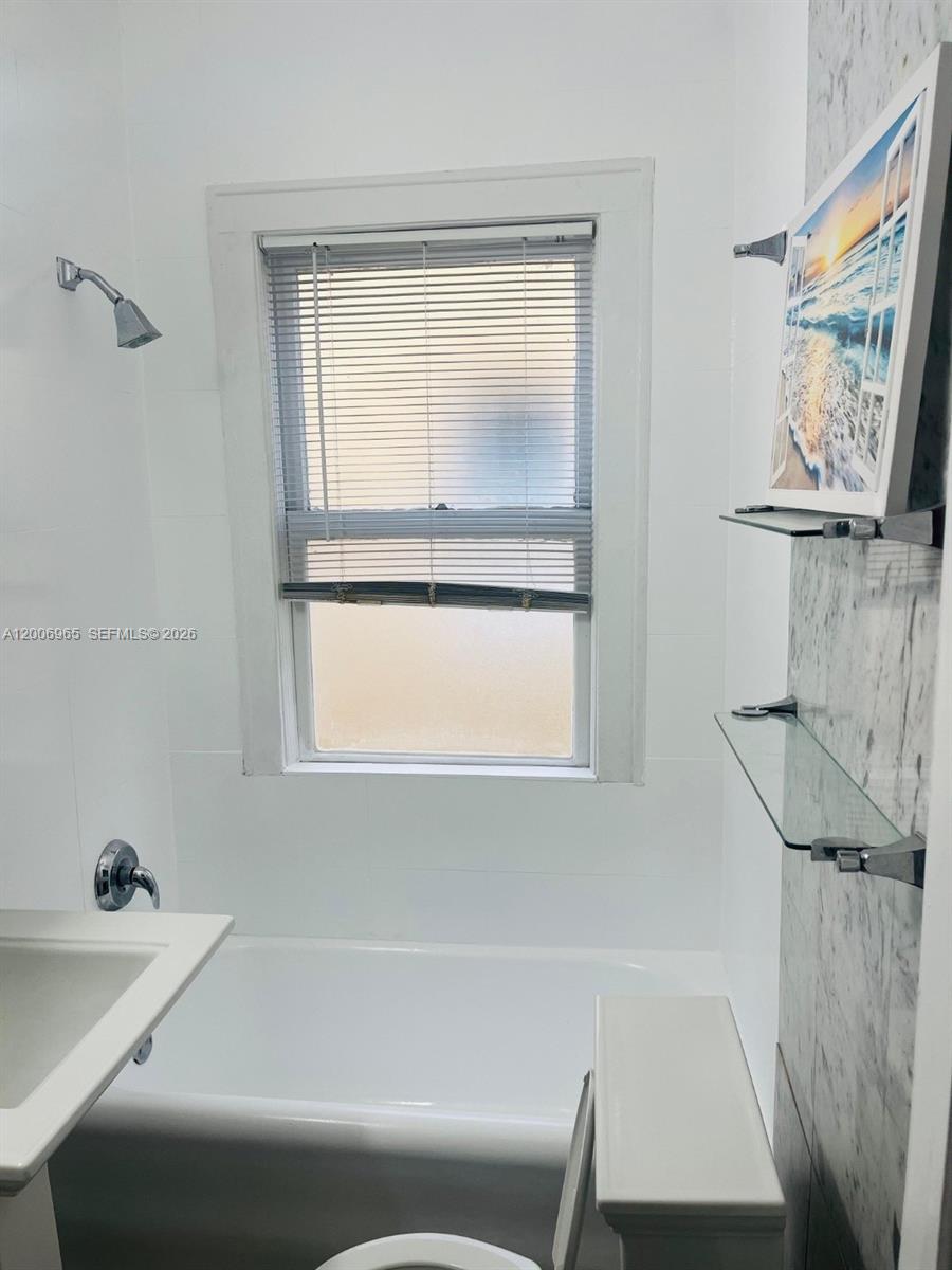 A bathroom features a white bathtub, a shower head, a window, and glass wall shelves — CORAL GABLES, FL